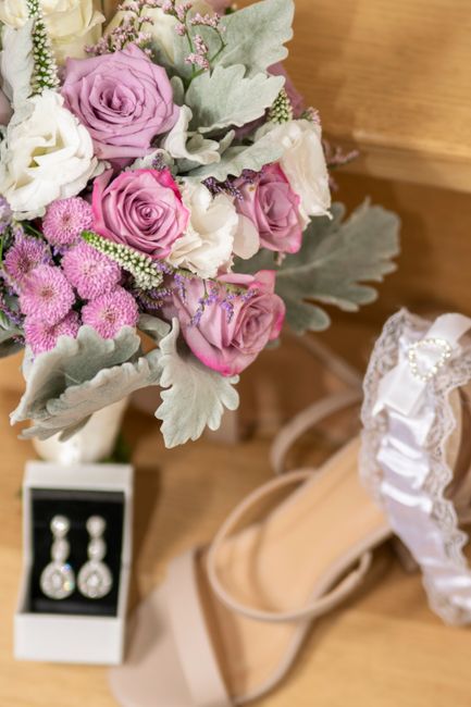 A close-up of a floral arrangement featuring soft pink roses, white lisianthus, and clusters of small pink asters, accented with silvery-grey dusty miller leaves, arranged in a bouquet style. The flow