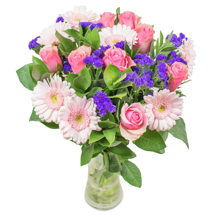Elegantly displayed in a clear glass vase positioned against a crisp white background, this Biggin Hill bouquet exudes both vitality and refined charm. At its heart, 7 to 9 pink roses stand out, each bloom displaying petals that gently curl outward or remain just partially open, highlighting their signature spirals and the promise of unfolding beauty. Interwoven with these are 5 to 7 pale pink gerbera daisies, their generous circular faces and whisper-soft petals radiating a calming pastel warmth, reminiscent of Biggin Hill's tranquil gardens in early summer. Clusters of vivid purple statice, reminiscent of wildflowers found along nearby green lanes, add both texture and a pop of deeper, royal colour-bringing a balanced energy without overwhelming the serene core of the arrangement. Lush, deep green foliage spills generously around the base, providing not only a sense of abundance and freshness but a fragrant undertone that hints at woodland air after gentle rain. Stems and water are visible through the vase, reinforcing the bouquet's freshness and the authenticity typical of a delivery from Florist Biggin Hill. Perfect for birthdays, anniversaries, Mother's Day, or simply to add a note of vibrant affection to a home or office, this composition speaks of celebration, care, and the simple pleasures of giving flowers in the heart of Biggin Hill.